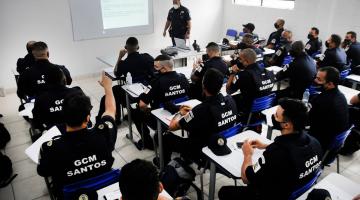 Guardas municipais uniformizados estão sentados em cadeira de sala de aula atentos a um homem que fala a todos, no fundo da classe. Na parede está projetada a imagem de um texto. #paratodosverem