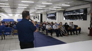 homem está falando ao microfone e à frente de uma mesa. Ele está de costas para a foto. Várias pessoas estão sentadas no auditório. #paratodosverem