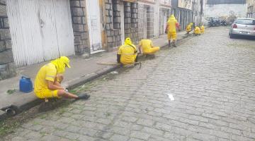 rua com sete homens sentados na calçada e um em pé retirando mato junto às sarjetas. #paratodosverem 