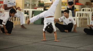 menina está dando estrela em aula de capoeira. Suas pernas estão abertas no ar. As duas mãos no chão. Ao fundo, mais crianças agachadas. 