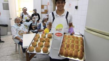 homem segura duas bandejás com pães de cará recém-saídos do forno. Atrás dele as alunas estão sentadas. #paratodosverem