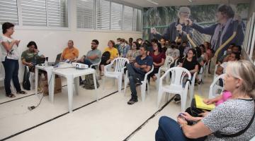 sala com pessoas sentadas e uma mulher em pé falando a todos. #paratodosverem