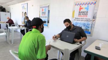 homem está sentado com laptop aberto à sua frente. Ele atende outro homem que está de costas para a foto. Ao fundo, um banner do banco do povo. #paratodosverem