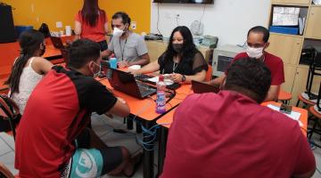 pessoas estão sentadas diante de mesa sendo atendidas por outras que estão diante de notebooks. #paratodosverem