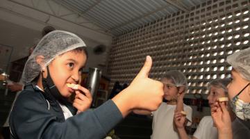 menina come pão com patê e faz sinal de positivo. Outras crianças estão no ambiente comendo o mesmo. #paratodosverem