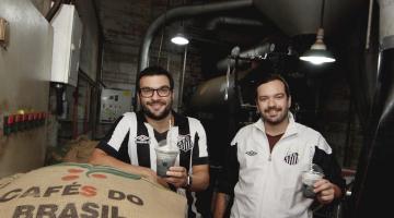 dois jovens com camisa do santos segurando café com saco de fé a frente e maquinário ao fundo 