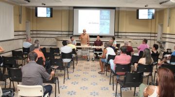 pessoas sentadas, de costas para a foto, assistindo a uma palestra. Ao fundo, uma mesa com três pessoas, sendo uma delas em pé. #paratodosverem 