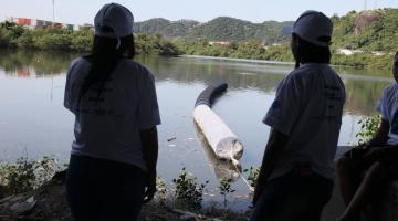 duas pessoas olham para trecho de mar com barreira flutuante. #paratodosverem 