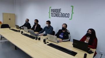 pessoas sentadas à mesa, com seus respectivos notebooks. Na parede está fixada a placa do parque tecnológico. #paratodosverem