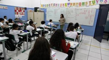 alunos estão em sala assistindo aula. Todos de costas para a foto e a professora em pé, ao fundo, de frente. #paratodosverem