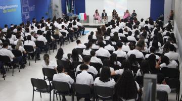 várias pessoas sentadas em auditório assistindo palestra com cinco pessoas no palco ao fundo. #paratodosverem