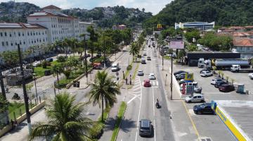 vista aérea de avenida com estabelecimentos de ambos os lados e um canteiro central com palmeiras. Carros cortam a via,. #paratodosverem 