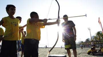 criança aponta arco e flecha na praia com professor olhando #paratodosverem