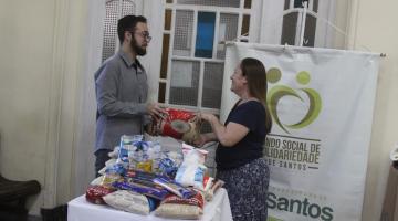 homem segura pacote de arroz ao mesmo tempo que mulher. ambos estão atrás de uma mesa com mantimentos e ao lado do banner do fundo social de solidariedade. #paratodoverem 