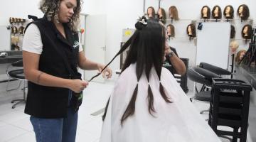 mulher está em pé cortando mechas de cabelo de uma mulher que está sentada. #paratodosverem