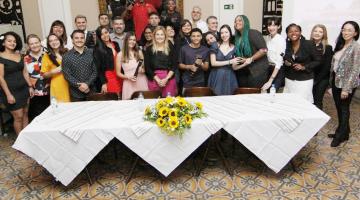 turma de alunos com dirigentes posa para foto atrás de uma mesa decorada com um buquê de flores. #paratodosverem