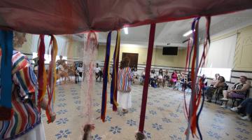 Tenda com fios coloridos em primeiro plano e mulheres sentadas em roda ao fundo. #paratodosverem 