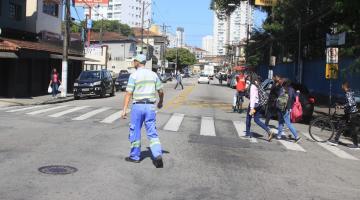 agente está no meio de uma rua, de costas para a foto e de frente para faixa de pedestres. Algumas pessoas cruzam a rua. #paratodosverem