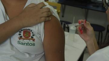 mão de mulher vacina braço de criança que usa uniforme da rede municipal. brasão de santos está no peito, à direita. criança segura a manga da camisa. #paratodosverem 