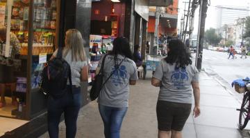 três mulheres com camiseta onde se lê nas costas amigos do bairro caminham por calçada. #paratodosverem