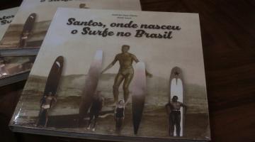 livros sofre a mesa. a foto é de alguns surfistas na praia com pranchões antigos. no alto se lê Santos, onde nasceu o surfe no Brasil. #paratodosverem 