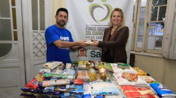 organizador do evento entrega alimento para representante do fundo com vários alimentos em mesa #paratodosverem
