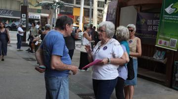 mulher segurando pracheta está conversando com homem. Ao fundo, há banner pendurado com informações para o segmento da terceira idade. #paratodosverem 