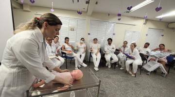 mulher massageia boneco diante de mulheres que estão sentadas assistindo. #paratodosverem 