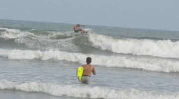 criança está entrando no mar com prancha em baixo do braço esquerdo (em primeiro plano) e outra criança surfando ao fundo. #paratodosverem 