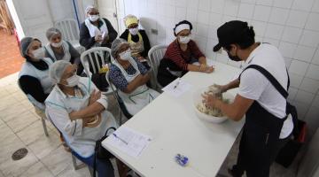 homem está sovando massa de pão na frente de alunas que estão sentadas. #paratodosverem