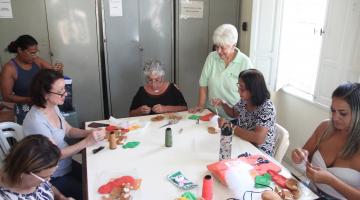 mulheres sentadas em torno de mesa fazem artesanato. #paratodosverem 