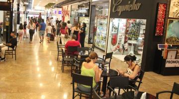 corredor de shopping com pessoas snetadas em mesas de cafezinho e circulando. #paratodosverem 