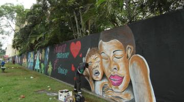 homem está grafitando muro. por trás, muitas árvores. #paratodosverem