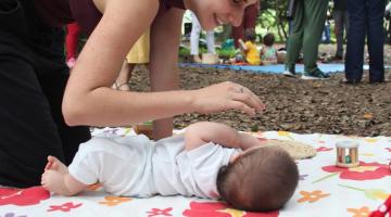 bebê está deitado no chão sobre colchonete. Mãe está ajoelhada olhando para ele e dirigindo uma das mãos ao rosto da criança. #paratodosverem 