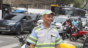Ricardo está na rua, próximo à via e a frente da moto que usa para trabalhar. Há carros parados no semáforo ao fundo. #paratodosverem