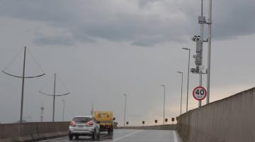 veículos trafegam em viaduto. Ao lado direito. Uma placa indicando o limite de velocidade e o radar instalado num poste. #paratodosverem
