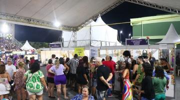 área de alimentacão com pessoas sentadas e pé próximas a mesas e barracas de lanches. #paratodosverem 