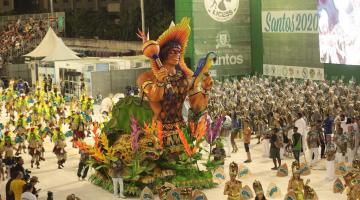 carro alegórico de indios e várias pessoas participando de desfile ao redor em passarela #paratodosverem