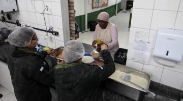 mulheres atrás de balcão servem comida no prato a uma outra mulher. #paratodosverem