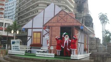 Papai e mamãe noel estão acenando na frente da casinha, no meio de uma praça. #psaratodosverem