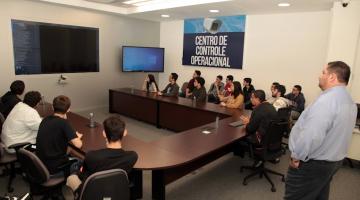 estudantes estão sentados em torno de uma mesa e diante de um telão. #paratodosverem