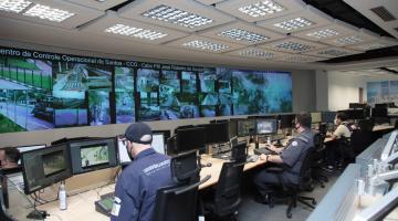 Guardas atuam no Centro de Controle Operacional. Eles estão diante de um telão onde são transmitidas imagens da Cidade. #Paratodosverem