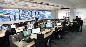 Sala do centro de controle operacional com telões e pessoas trabalhando diante de monitores #Paratodosverem