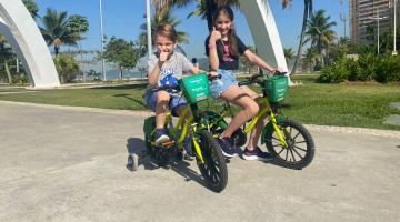 duas crianças estão pedalando em bicicletas infantis com rodinhas. Elas fazem sinal positivo com as respectivas mãos direitas.#paratodosverem 