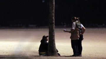 Agentes abordam pessoa em situação de rua na faixa de areia da praia, próximo a uma palmeira. A ação é realizada à noite. #Paratodosverem