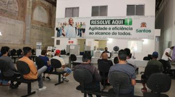 pessoas estão sentadas em grande salão. Ao fundo e no alto, afixada na parede, há uma grande placa onde se lê Resolve Aqui - agilidade e eficiência ao alcance de todos. O logotipo da prefeitura aparece à direita. #paratodosverem