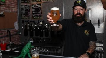 homem estende a mão e o braço com copo de cerveja cheio, como se estivesse oferecendo para a foto. #paratodosverem