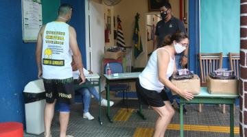 Mulher suspende cesta básica que está sobre uma mesa. Ao lado, um homem aguarda para ser atendido. #Paratodosverem