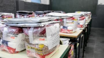 pacotes de cestas básicas estão sobre mesas escolares. #paratodosverem
