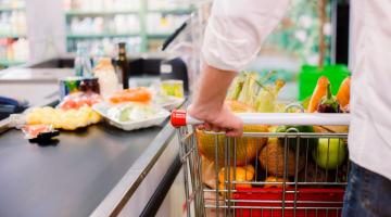 carrinho com alimentos junto à caixa de supermercado. Mãos masculinas seguram no veículo. #paratodosverem 